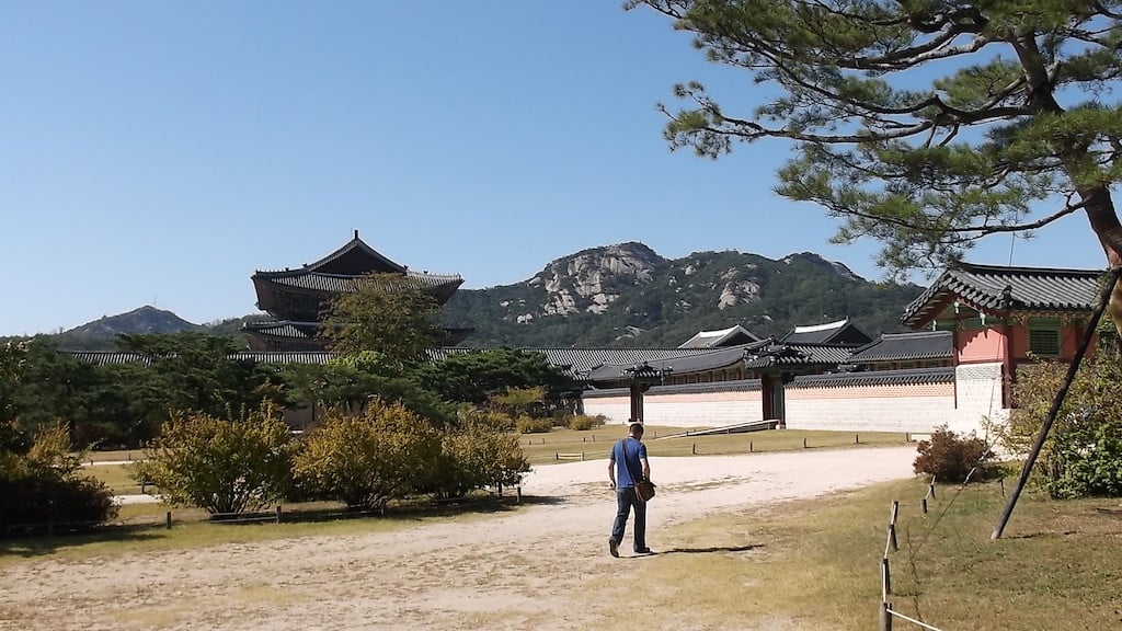 ALT: A landscape photograph with a mountain ridge in the background. The foreground has some scattered shrubs on dusty grass. In front of the mountains sits a series of red and white hanoks - a type of traditional South Korean architecture with sloping tiled roofs.